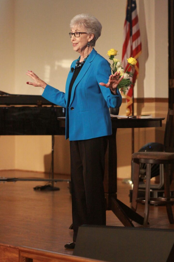 An older woman standing on stage in front of a piano.